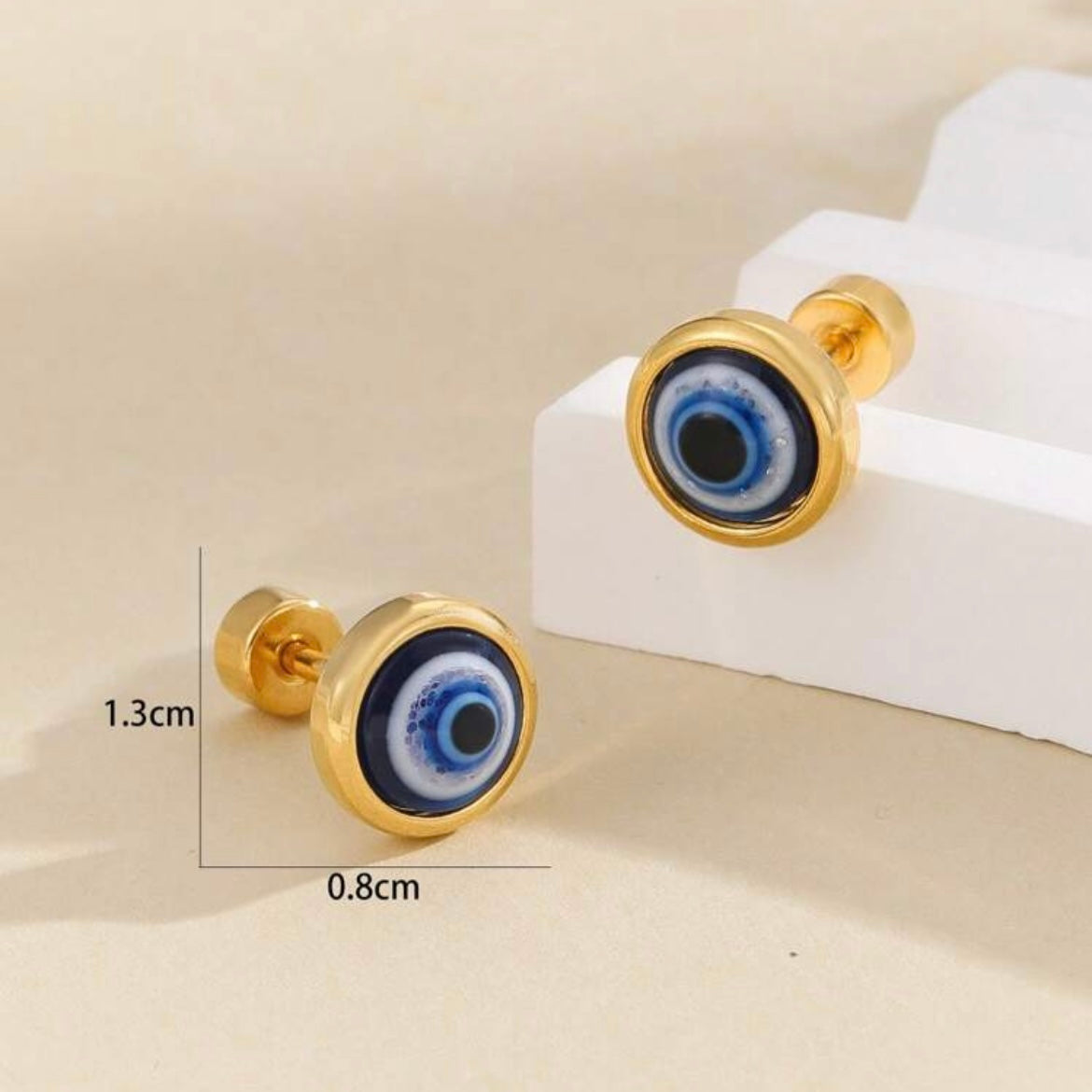 Gold earrings with blue eye design on a beige surface, showing measurements.